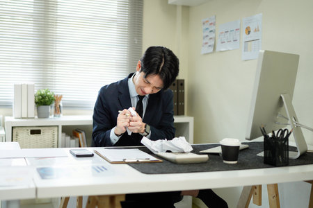 An asian businessman looks stressed while working at his desk, with crumpled paper surrounding him, symbolizing writer's block..の写真素材