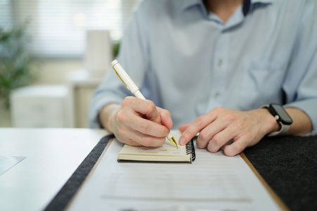 Businessman analyzing data (graphs/charts) on a document, taking notes with a pen. Office setting, professional work.の写真素材
