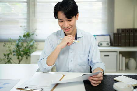 Smiling young executive analyzing business documents. Male professional is working on laptop. He is wearing formals while sitting at office desk..の写真素材
