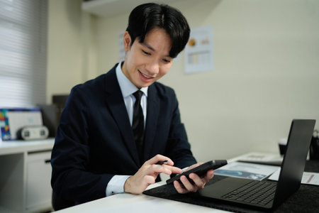 A man in a suit is using a cell phone while sitting at a desk. Concept of professionalism and focus, as the man is likely working on important tasks or communicating with colleagues.の写真素材
