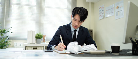 An asian businessman looks stressed while working at his desk, with crumpled paper surrounding him, symbolizing writer's block..の写真素材