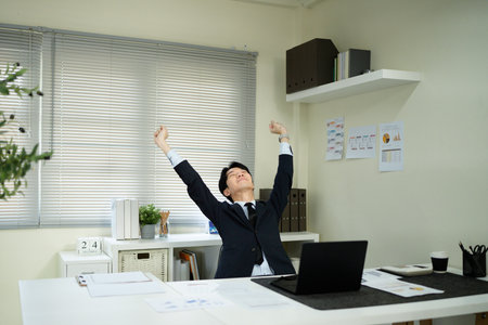A man in a suit is sitting at a desk with a laptop and a computer monitor. He is holding his arms up in the air, as if he is celebrating or expressing joy. Concept of accomplishment or satisfactionの写真素材