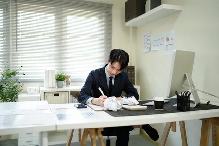 An asian businessman looks stressed while working at his desk, with crumpled paper surrounding him, symbolizing writer's block..の写真素材