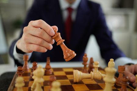 Person suit holding wood chess king piece above chessboard with other yellow chess pieces focus, showing strategic thinking and concentration.の写真素材