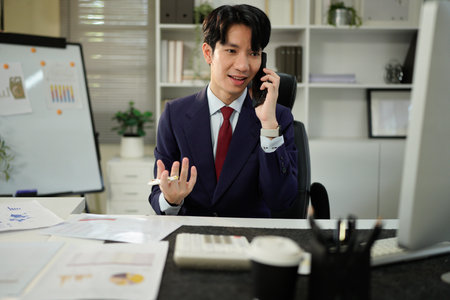 Image of handsome businessman talking on cell phone while working on computer in office.の写真素材