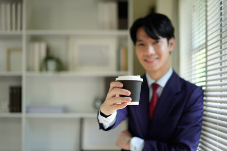 Happy young businessman in suit smiling and holding a coffee cup near window He is enjoying a morning beverage.の写真素材