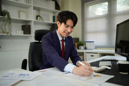 Young man reading documents at desk in modern office with computer and bookshelf nearby, showcasing a professional work environment under soft natural light..の写真素材