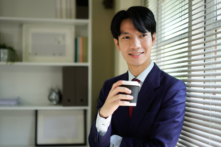 Happy young businessman in suit smiling and holding a coffee cup near window He is enjoying a morning beverage.の写真素材