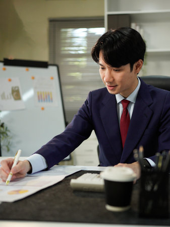 Businessman analyzing data (graphs/charts) on a document, taking notes with a pen. Office setting, professional work.の写真素材
