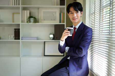 Happy young businessman in suit smiling and holding a coffee cup near window He is enjoying a morning beverage.の写真素材