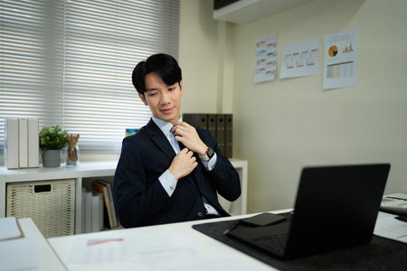 A man in a suit is sitting at a desk with a laptop in front of him. He is wearing a tie and he is focused on his work. Concept of professionalism and productivityの写真素材