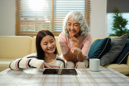 Smiling granddaughter and grandmother with tablet pc computer sitting on couch at home..の写真素材