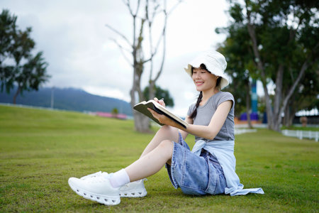 woman, writing and book in park, thinking and relax by tree, grass or sunshine for peace. Girl, person or student with story in nature, college or notes with vision, knowledge or campus lawn.の写真素材