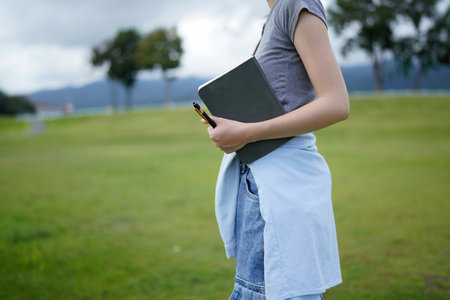 A woman is holding a book and a pen in a park. The book is a notebook. The scene is peaceful and serene, with the woman standing in a grassy fieldの写真素材