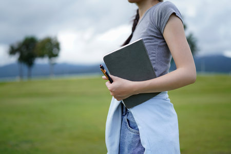 A woman is holding a book and a pen in a park. The book is a notebook. The scene is peaceful and serene, with the woman standing in a grassy field.の写真素材