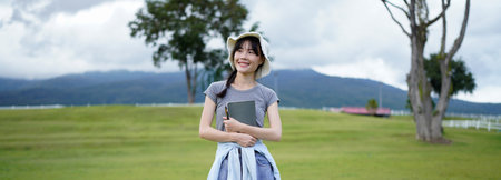 A woman is holding a book and a pen in a park. The book is a notebook. The scene is peaceful and serene, with the woman standing in a grassy field.の写真素材