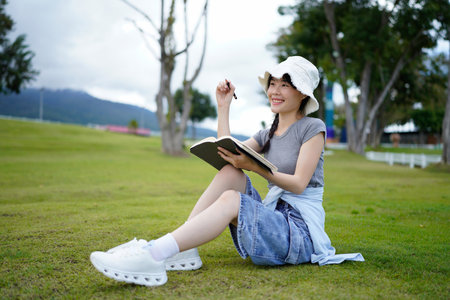 woman, writing and book in park, thinking and relax by tree, grass or sunshine for peace. Girl, person or student with story in nature, college or notes with vision, knowledge or campus lawn.の写真素材