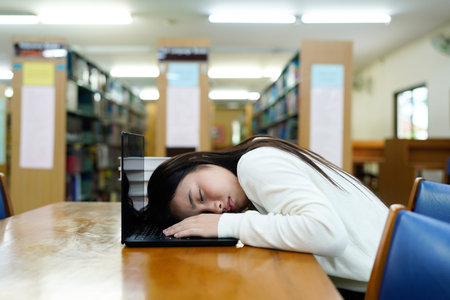 A tired student asleep at a desk in a library, with a laptop and books nearby..