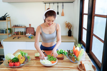 Happy asian fitness woman listening to music with headphones and preparing a vegetable salad in the kitchen, enjoying a healthy lifestyle.の写真素材