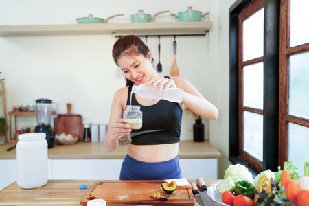 Smiling young woman in sports clothing cooking protein cocktail while standing at the home kitchen Healthy lifestyle concept.の写真素材