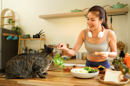 Asian woman in sportswear preparing salad in a kitchen and feeding a tabby cat.の写真素材