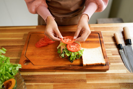 woman prepares a sandwich at home, layering ham and fresh vegetables in a warm, natural light...
