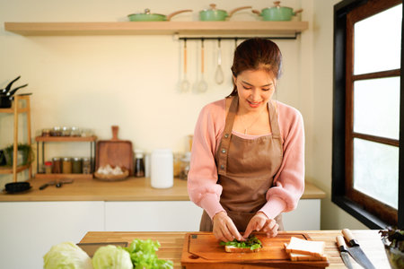 woman prepares a sandwich at home, layering ham and fresh vegetables in a warm, natural light kitchen.の写真素材