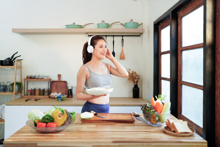 Happy asian fitness woman listening to music with headphones and preparing a vegetable salad in the kitchen, enjoying a healthy lifestyle.の写真素材