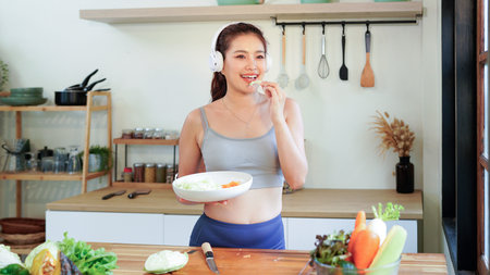 Happy asian fitness woman listening to music with headphones and preparing a vegetable salad in the...