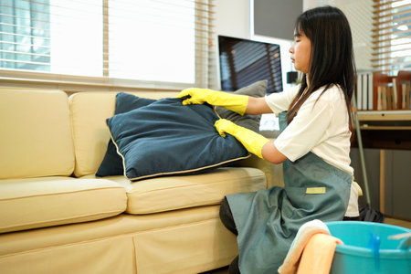 Housekeeper wearing yellow gloves is arranging pillows on a sofa in the living room, ensuring a tidy and comfortable environment for residents or guests.の写真素材