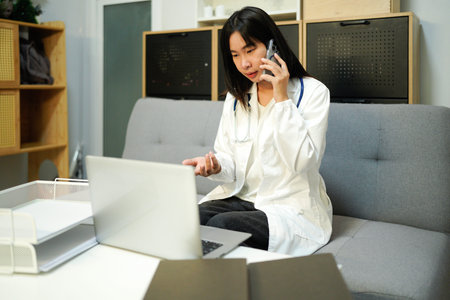 Cheerful young female doctor in uniform sitting at workplace at desk in office talking by mobile phone and working on a laptop computer in medical clinic. Health care and medicine concept.の写真素材