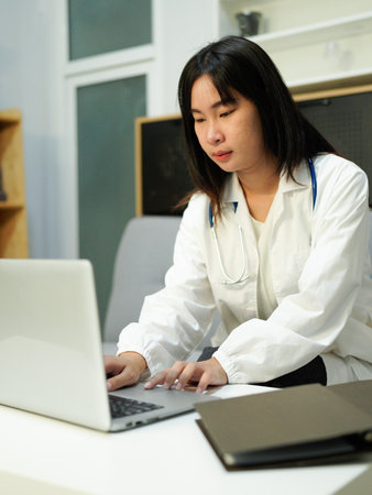 Cropped photo of female doctor in white uniform working on laptop computer in hospital, sitting at desk with stethoscope and ultrasound results.の写真素材