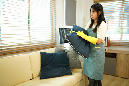 Housekeeper wearing yellow gloves is arranging pillows on a sofa in the living room, ensuring a tidy...