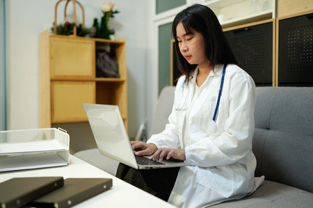 Cropped photo of female doctor in white uniform working on laptop computer in hospital, sitting at desk with stethoscope and ultrasound results.の写真素材