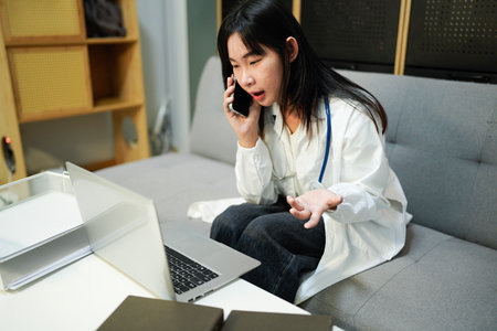 Cheerful young female doctor in uniform sitting at workplace at desk in office talking by mobile phone and working on a laptop computer in medical clinic. Health care and medicine concept.の写真素材