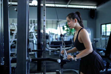 Fit Asian woman pulling down weights on a cable machine at the gym, training her biceps and building...