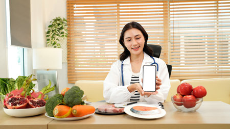 Young Asian female nutritionist in a white lab coat, working at a desk. She shows smartphone with blank screen consultation details, promoting weight loss and balanced diets.の写真素材