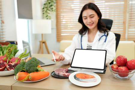 Young Asian female nutritionist in a white lab coat, working at a desk. She shows tablet with blank screen consultation details, promoting weight loss and balanced diets.の写真素材