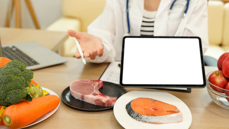 Young Asian female nutritionist in a white lab coat, working at a desk. She shows tablet with blank screen consultation details, promoting weight loss and balanced diets.の写真素材