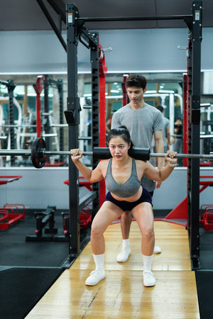 Professional fitness trainer guiding female athlete performing precise barbell squats in modern gym...
