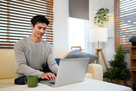 Young asian male tech user relaxing holding laptop computer and looking at the screen in living room, Remote Job or work from home concept..の写真素材