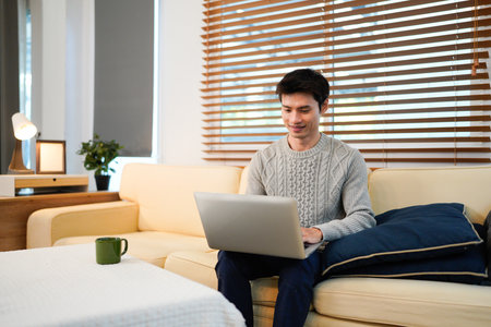 Young asian male tech user relaxing holding laptop computer and looking at the screen in living room, Remote Job or work from home concept..の写真素材
