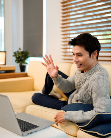Confident Asian man using laptop while lay down on cozy sofa at modern home.