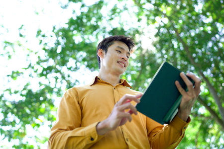 Man enjoys reading a book under a large tree in a serene park on a beautiful sunny day.の写真素材