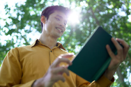 Man enjoys reading a book under a large tree in a serene park on a beautiful sunny day.の写真素材