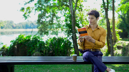 young man enjoying leisure time on a park bench, reading a book and drinking coffee on a sunny day...