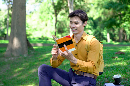 Man enjoys reading a book under a large tree in a serene park on a beautiful sunny day.