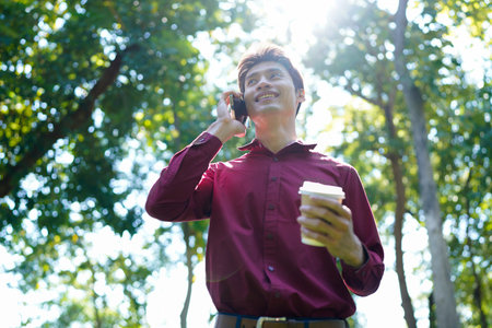 Young asian man with takeout coffee communicating on smartphone at urban park, blank space.