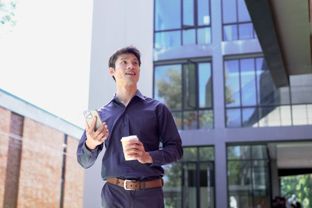Smiling businessman using smartphone and holding coffee outside office building.