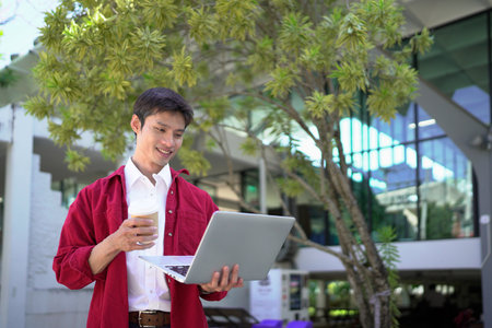Man working remotely outdoors, drinking coffee and using laptop in city park.